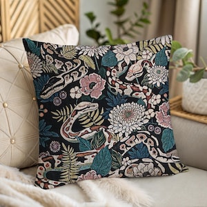 Peut inclure: Coussin carré décoratif avec un fond noir orné d'un motif de serpents, de fleurs et de feuillage blancs, roses, verts et bleus. Le coussin est posé sur un canapé beige avec une couverture crème.