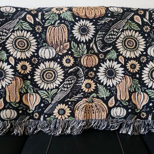 May include: Black throw blanket with a repeating pattern of sunflowers, pumpkins, and crows. The blanket has a fringed edge and is made of a soft, woven material. The color palette includes shades of black, beige, orange, and green.