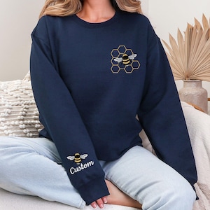 May include: A navy blue sweatshirt with a bee and honeycomb design on the chest and a bee design on the left sleeve. The text "Custom" is printed below the bee on the sleeve.