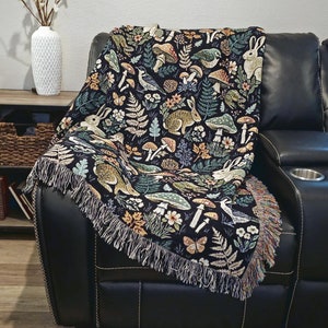 May include: A black throw blanket with a forest-themed pattern featuring rabbits, mushrooms, and foliage in shades of green, brown, and cream. The blanket has a fringed edge and is draped over a black leather recliner.