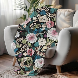 Może przedstawiać: Czarny koc z kwiatowym i wężowym wzorem, narzucony na biały fotel. Koc ma różowe, białe i niebieskie kwiaty, zielone liście oraz brązowe i białe węże.