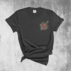 Puede incluir: Una camiseta negra con un dise&ntilde;o de tatuaje de estilo vintage de una daga y rosas en el pecho izquierdo.