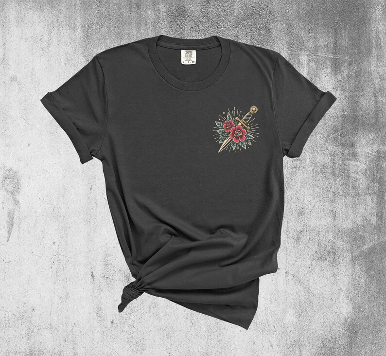 Puede incluir: Una camiseta negra con un dise&ntilde;o de tatuaje de estilo vintage de una daga y rosas en el pecho izquierdo.