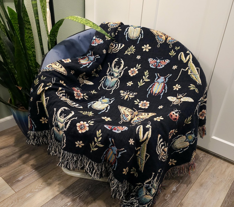 May include: A black woven throw blanket with a fringe border features a repeating pattern of colorful insects, butterflies, and flowers.