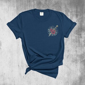 Puede incluir: Una camiseta azul marino con un dise&ntilde;o de tatuaje vintage de una daga y rosas rojas en el pecho izquierdo.