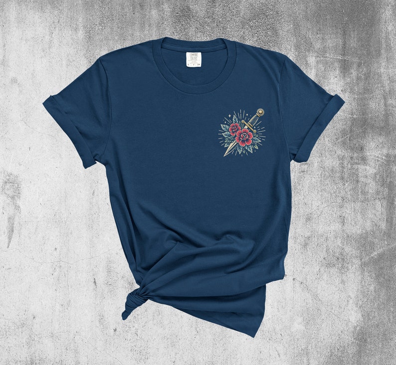 Puede incluir: Una camiseta azul marino con un dise&ntilde;o de tatuaje vintage de una daga y rosas rojas en el pecho izquierdo.