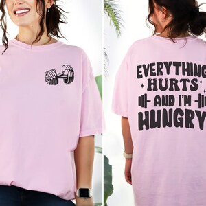 May include: Pink t-shirt with a dumbbell graphic on the front and the text "Everything Hurts And I'm Hungry" on the back.