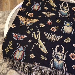 May include: Black woven throw blanket with a repeating pattern of colorful butterflies, beetles, and praying mantises. The blanket has a fringe border.