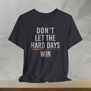 Puede incluir: Una camiseta negra con el texto "DON'T LET THE HARD DAYS WIN" impreso en blanco. Una l&iacute;nea roja se dibuja a trav&eacute;s de la palabra "HARD".