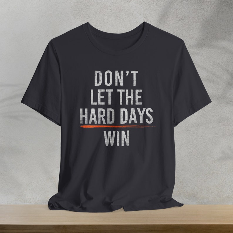 Puede incluir: Una camiseta negra con el texto "DON'T LET THE HARD DAYS WIN" impreso en blanco. Una l&iacute;nea roja se dibuja a trav&eacute;s de la palabra "HARD".