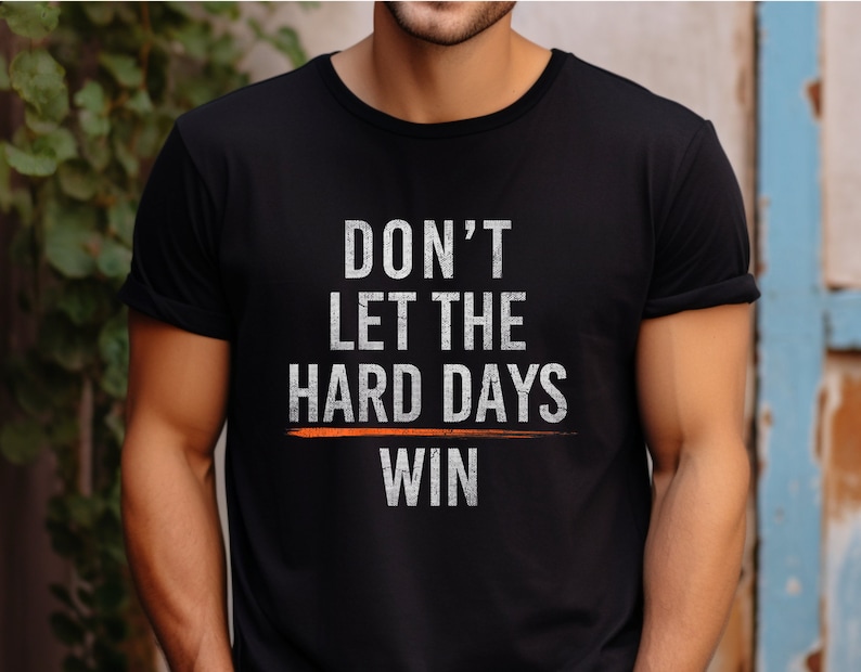 Puede incluir: Camiseta negra con un gr&aacute;fico blanco que dice "No dejes que los d&iacute;as dif&iacute;ciles ganen". El gr&aacute;fico tiene una l&iacute;nea naranja horizontal que lo atraviesa.