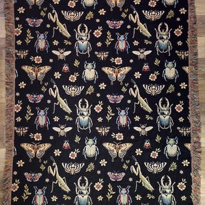 May include: Black woven throw blanket with a repeating pattern of butterflies, beetles, praying mantises, and flowers. The pattern is in shades of blue, green, red, and yellow.