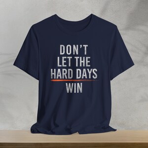 Puede incluir: Camiseta azul marino con un gr&aacute;fico blanco que dice "DON'T LET THE HARD DAYS WIN" con una l&iacute;nea roja debajo de "HARD DAYS".