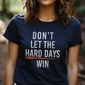 Puede incluir: Una camiseta azul marino con un gr&aacute;fico blanco que dice "No dejes que los d&iacute;as dif&iacute;ciles ganen". El gr&aacute;fico tiene una l&iacute;nea blanca a trav&eacute;s de la palabra "dif&iacute;ciles" y una l&iacute;nea naranja a trav&eacute;s de la palabra "ganar".