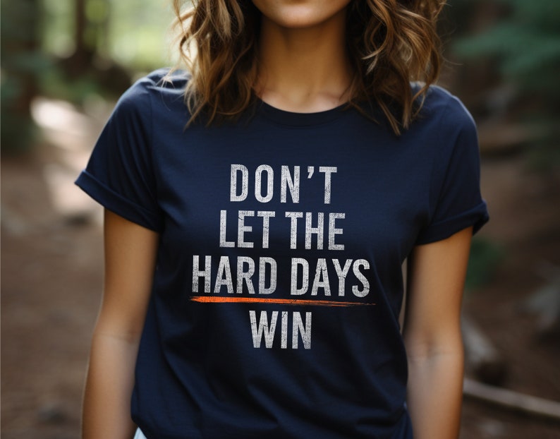 Puede incluir: Una camiseta azul marino con un gr&aacute;fico blanco que dice "No dejes que los d&iacute;as dif&iacute;ciles ganen". El gr&aacute;fico tiene una l&iacute;nea blanca a trav&eacute;s de la palabra "dif&iacute;ciles" y una l&iacute;nea naranja a trav&eacute;s de la palabra "ganar".