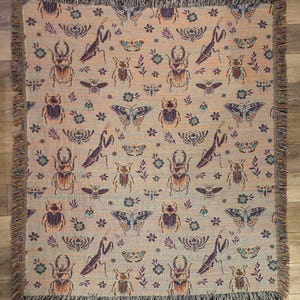 May include: A woven throw blanket with a beige background and a repeating pattern of butterflies, beetles, and praying mantises in shades of purple, green, and brown.