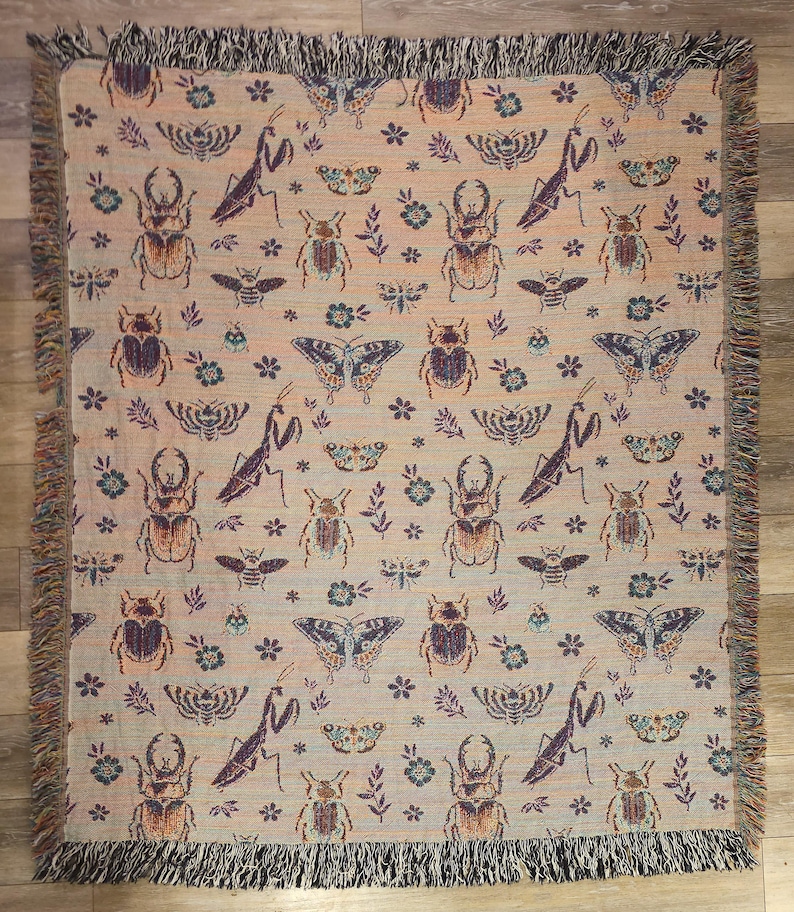 May include: A woven throw blanket with a beige background and a repeating pattern of butterflies, beetles, and praying mantises in shades of purple, green, and brown.