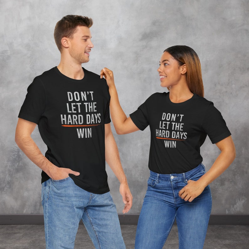 Puede incluir: Dos personas con camisetas negras con el texto "Don't let the hard days win" en blanco y naranja. Las camisetas son un mensaje motivacional.