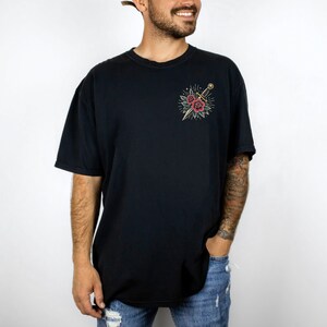 Puede incluir: Una camiseta negra con un gr&aacute;fico de una daga y flores. El gr&aacute;fico est&aacute; en un estilo vintage.