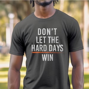 Puede incluir: Una camiseta gris oscuro con el texto "DON'T LET THE HARD DAYS WIN" en blanco con una l&iacute;nea roja debajo de la palabra "DAYS".