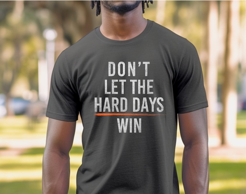 Puede incluir: Una camiseta gris oscuro con el texto "DON'T LET THE HARD DAYS WIN" en blanco con una l&iacute;nea roja debajo de la palabra "DAYS".