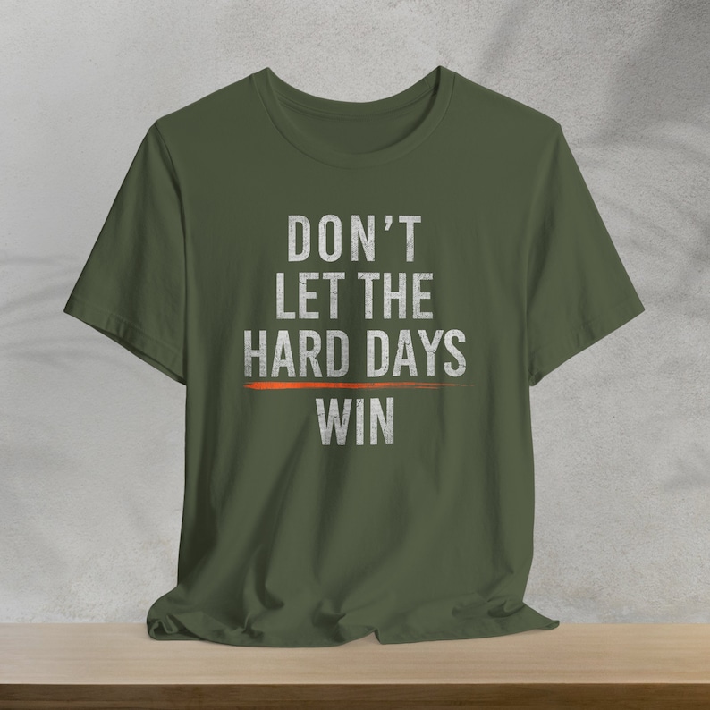 Puede incluir: Una camiseta verde con el texto "DON'T LET THE HARD DAYS WIN" en blanco con una l&iacute;nea roja debajo de "HARD DAYS".