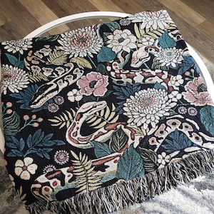 May include: Black woven throw blanket with a floral and snake pattern. The design features white, pink, and teal flowers, along with detailed snake illustrations. The blanket has a fringed edge.