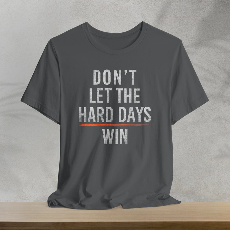 Puede incluir: Una camiseta gris oscuro con el texto "DON'T LET THE HARD DAYS WIN" impreso en blanco. Una l&iacute;nea roja se dibuja debajo de la palabra "WIN".