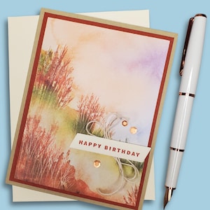 Op de afbeelding: Een verjaardagskaart met een aquarel landschapsontwerp in tinten oranje, groen en paars. De kaart heeft de tekst "HAPPY BIRTHDAY" en is versierd met koperkleurige accenten. Een witte pen met gouden accenten ligt naast de kaart.