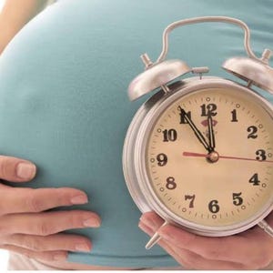 以下が含まれることがあります： 白い文字盤と黒い数字の銀色の目覚まし時計が12:00に設定されています。時計は妊娠中の女性が持っています。