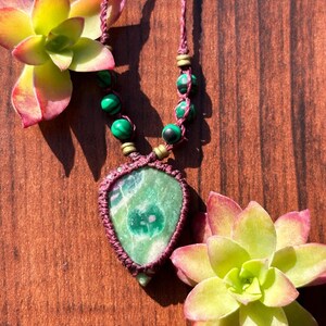 Può includere: Una collana fatta a mano con un ciondolo in malachite verde con un'incastonatura in macramè bordeaux. La collana include perline verdi e nere e un cordino bordeaux. Due piante grasse con foglie gialle e rosse incorniciano la collana.