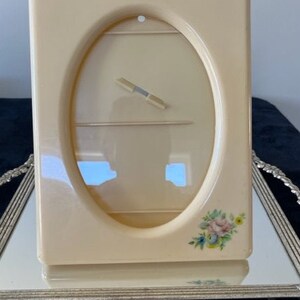May include: A rectangular cream-colored picture frame with a central oval opening and a clear protective cover. The frame features a small, delicate floral decal in the bottom right corner, showcasing pink, blue, and yellow blossoms.