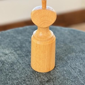 May include: A light-colored wooden pepper mill with a rounded top and cylindrical base. The pepper mill has a small, vertical handle on top and several small holes for dispensing pepper. The wood grain is visible.
