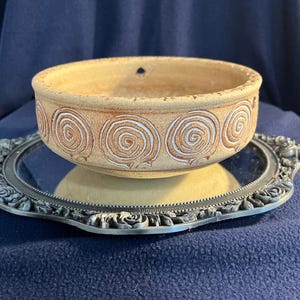 May include: A beige ceramic hanging planter with a white swirl design. The planter has a hole for hanging and is sitting on a silver tray with a decorative edge.