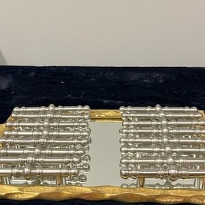 May include: A rectangular serving tray with a textured gold-tone frame and two side handles. The center features a mirrored surface topped with two rows of parallel silver-tone metal bars, each with a decorative, rounded, multi-segmented design.