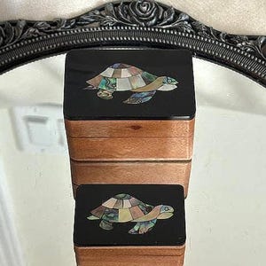 May include: Two wooden boxes stacked on top of each other. The boxes have a black top with a turtle design made of inlaid iridescent shell pieces. The sides of the boxes are a light brown color. The boxes are sitting on a reflective surface.