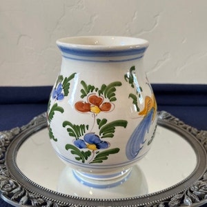 May include: A white ceramic vase with a blue rim and floral designs in shades of blue, green, yellow, orange, and red. The vase is sitting on a silver tray with a mirrored surface.
