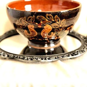 May include: A brown ceramic bowl with a decorative design of stylized leaves and flowers in gold and orange. The bowl has a wide rim and a rounded base.