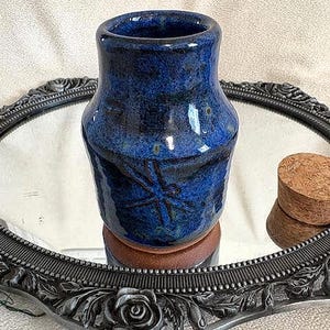 May include: A handmade, blue ceramic vase with a textured surface and a wide opening. The vase sits on a small, round, brown base and is next to a cork. The vase is displayed on a decorative, oval mirror with an ornate, silver-colored frame.