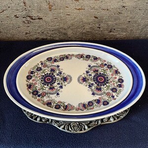 May include: A white oval serving platter with a blue rim and a floral design in shades of pink, blue, and green. The platter is sitting on a silver stand with a decorative edge.