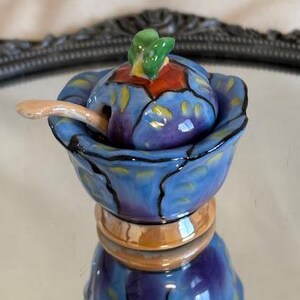 May include: A small blue and purple ceramic honey pot with a lid and a small spoon. The lid has a green leaf design and the pot has a floral pattern.