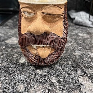 May include: A ceramic mug shaped like a face with a beard and tongue sticking out. The mug is tan with brown details for the beard and facial features. The mug is sitting on a speckled countertop.