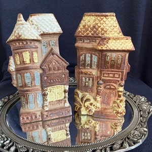 May include: Two ceramic houses with brown, yellow, and blue paint. The houses are detailed with windows, doors, and roofs. The houses are sitting on a silver tray with a floral design.