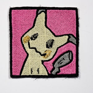 May include: An embroidered iron-on patch featuring a cartoon of a light yellow and brown Pokemon character with a gray tail, against a pink background.