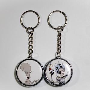 Op de afbeelding: Twee ronde sleutelhangers met zilveren kettingen en sleutelringen. Eén sleutelhanger toont een grijze alien die wijst, de andere een stripfiguur met een helm en schedels. Op een witte achtergrond.