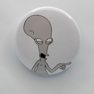 May include: A white button with a cartoon image of a gray alien with a large head and a long, thin body. The alien is pointing to the right with a finger and has a surprised expression on its face.