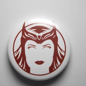 May include: A red and white button featuring a stylised illustration of a woman with a crown. The woman's eyes are closed and she has a red lip.