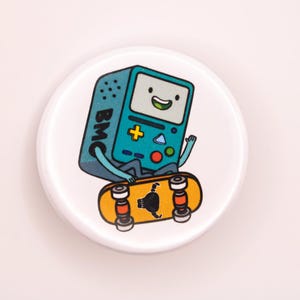 May include: A white round button featuring a cartoon character on a skateboard. The character is a blue and green video game console with a smiling face and the letters "BMC" on the side.