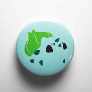 Peut inclure: Un bouton bleu et vert avec une image de dessin animé d'une créature aux cheveux verts et aux yeux noirs. La créature est entourée de petits points noirs.