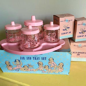 Puede incluir: Un juego de cuidado para bebés vintage con una bandeja rosa, frascos de vidrio con tapas y cajas etiquetadas como "Clean-Up Jar" y "Cotton Applicator Jar". El juego incluye una caja azul con ilustraciones de bebés.
